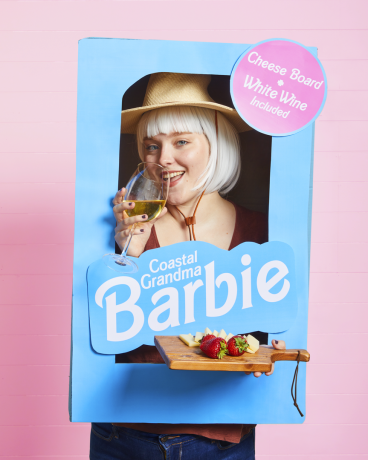 Déguisement de barbie de grand-mère côtière représentant une femme dans une boîte de barbie en carton bleu tenant une assiette de fromages et un verre de vin blanc