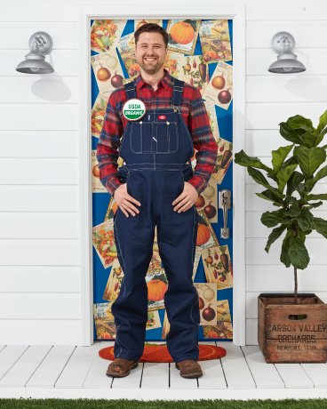 homme portant un costume d'agriculteur avec une salopette, une chemise à carreaux, un bouton biologique usda debout devant la porte décorée pour halloween