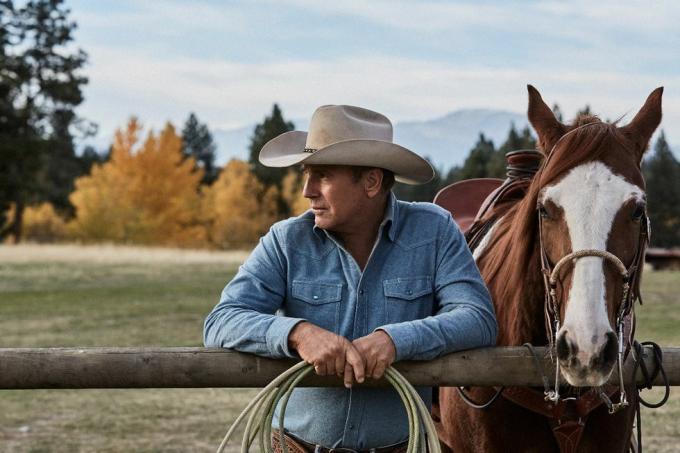 Kevin Costner à Yellowstone à côté d'un cheval appuyé sur une clôture avec une corde dans les mains portant une chemise en jean bleu délavé et un chapeau de cowboy beige
