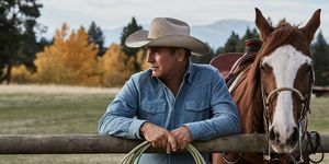 Kevin Costner à Yellowstone à côté d'un cheval appuyé sur une clôture avec une corde dans les mains portant une chemise en jean bleu délavé et un chapeau de cowboy beige