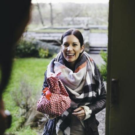 une femme sourit alors qu'elle se tient sur le pas de la porte pour rendre visite à un ami elle est enveloppée dans des vêtements chauds et tient un cadeau enveloppé dans un tissu à carreaux rouge et blanc