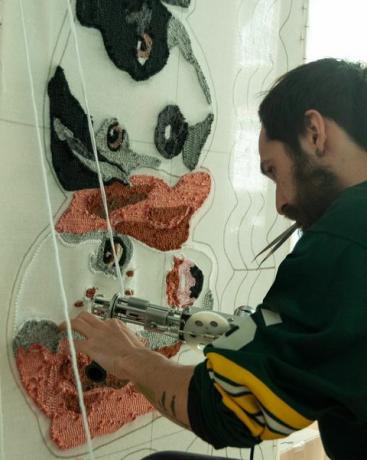 Homme barbu hispano-latino de 30 ans portant un maillot de rugby, travaillant dans son atelier sur des tapis artisanaux fabriqués à partir de tufting gun et de fibres acryliques, avec un design de tapis pour chien