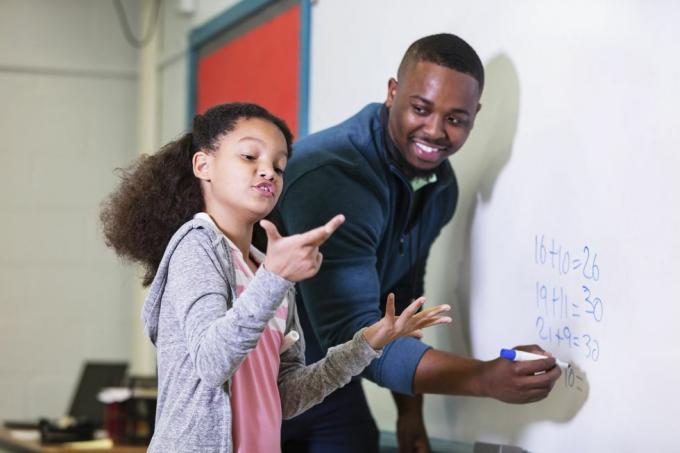 une fillette multiraciale de 9 ans en cours de mathématiques, debout au tableau blanc avec son professeur, un jeune afro-américain dans son 20 ans, l'enseignant regarde son élève du primaire, attendant qu'elle essaie de trouver la réponse, l'accent est mis sur la fille