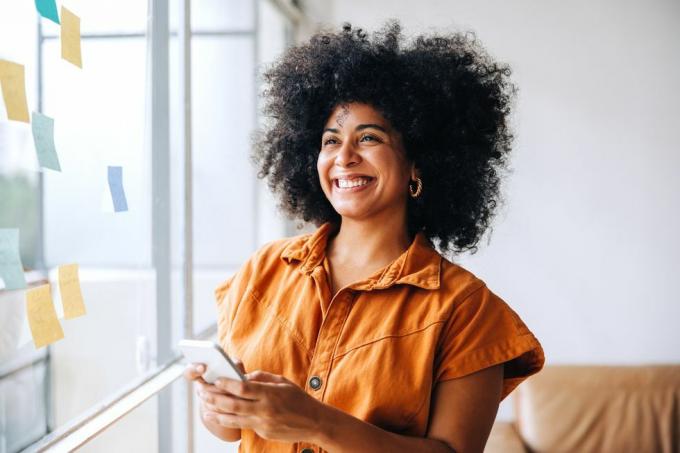 femme d'affaires noire heureuse souriant tout en utilisant un smartphone dans un bureau créatif femme entrepreneur joyeuse envoyant un message texte tout en se tenant à côté d'un mur de verre avec des notes autocollantes