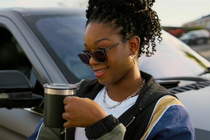 le personnage d'izzy se tient à côté d'un suv lincoln buvant une tasse de café, portant des lunettes de soleil