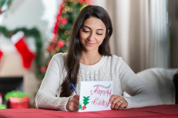 une jeune femme souriante est assise à une table dans son salon décoré et regarde avec concentration pendant qu'elle écrit une lettre à un ami pour Noël